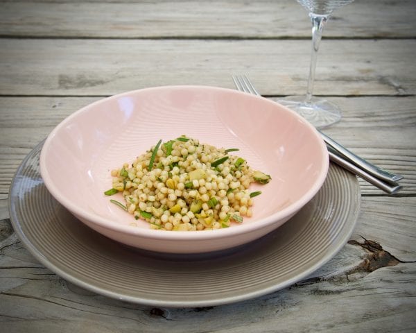 Foto van een kleurrijke Fregola Sarda salade met groene olijven, fijngehakte kruiden en citroenrasp, geserveerd in een grote schaal, perfect als bijgerecht bij gebraden lamskoteletjes.